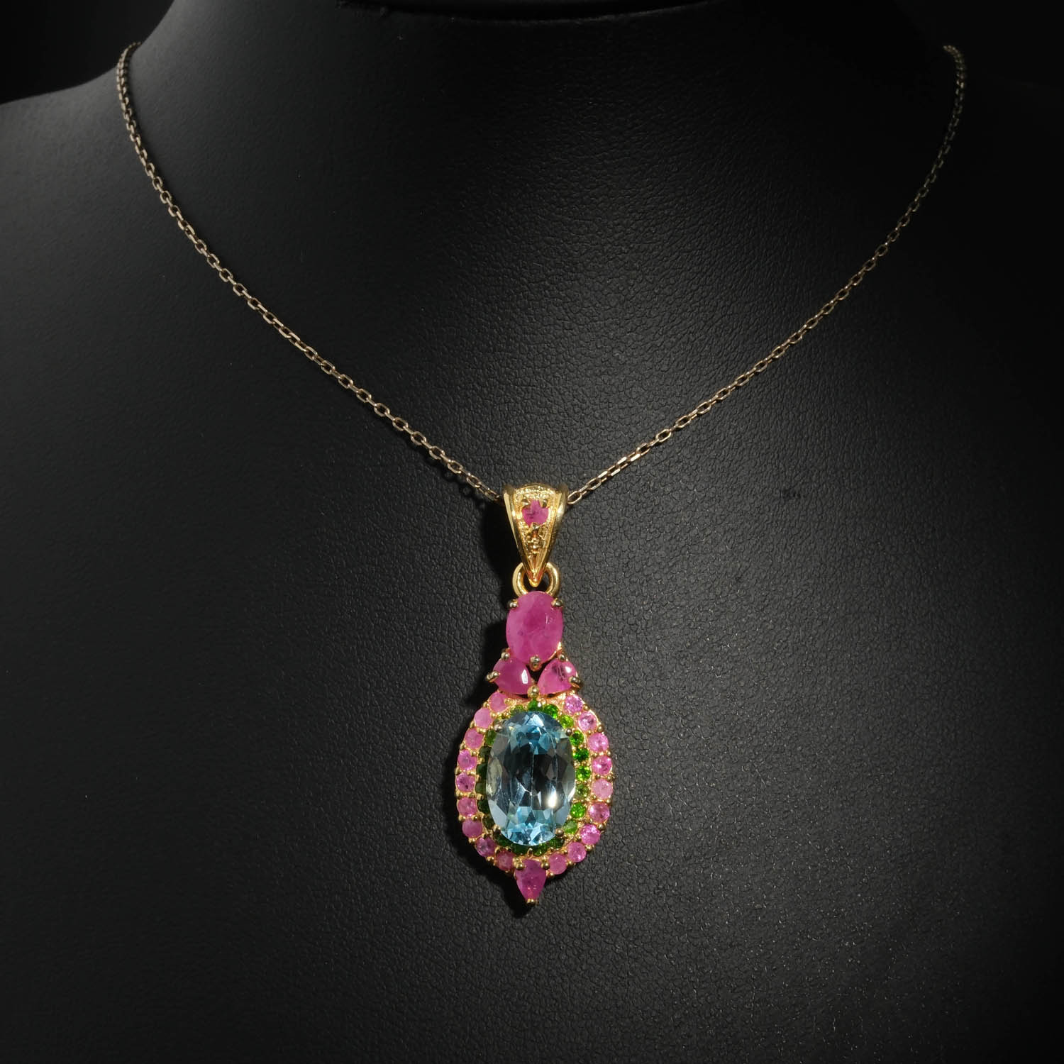 natural-blue-topaz-ruby-tsavorite-gold-plated-silver-necklace.jpg Natural faceted oval blue topaz pendant necklace with green tsavorite and ruby halo in gold plated 925 silver on white satin.