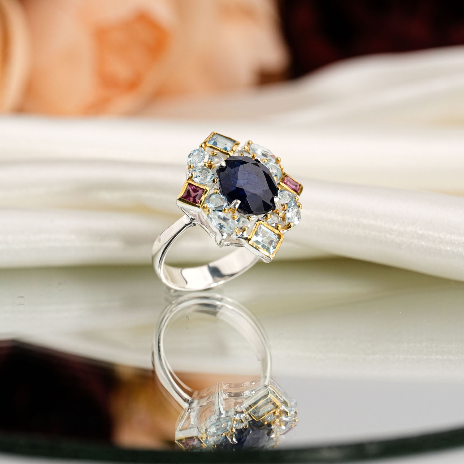 orijinal-faset-safir-blue-topaz-turmalin-925-gumus-kadin-yuzuk.jpg Beyaz saten kumaş ve ayna üzerinde faset oval lacivert safir etrafında açık mavi topaz ve pembe turmalin taşlı altın vurgulu 925 gümüş kadın yüzük.